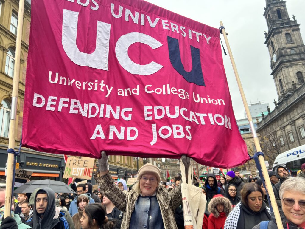 Megan Povey smiling and holding the UCU University of Leeds branch banner at a demonstration in Leeds opposing Israel's war in Gaza
